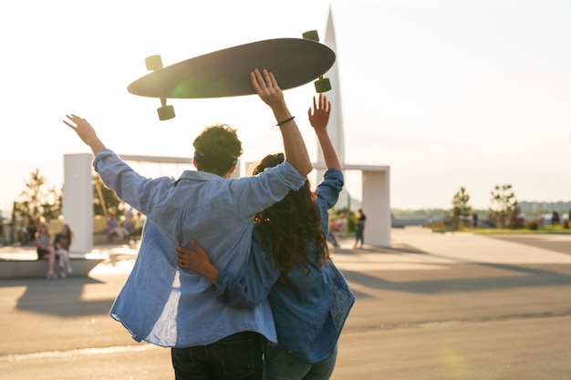 Glückliches Paar genießt die Umarmung des Sonnenuntergangs mit erhobenen Armen und hält das Longboard-Glück und das Freiheitskonzept