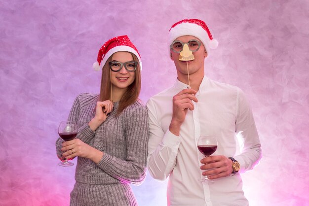 Glückliches Paar in Weihnachtsmützen mit Wein in Gläsern, die Maske in der Nähe des Gesichts halten und den Valentinstag feiern