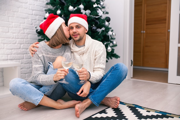 Glückliches Paar in Weihnachtsmützen nahe einem Weihnachtsbaum, der küsst