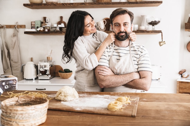 glückliches Paar Mann und Frau mit Schürzen, die zusammen Spaß haben, während sie hausgemachte Pasta in der Küche zu Hause zubereiten
