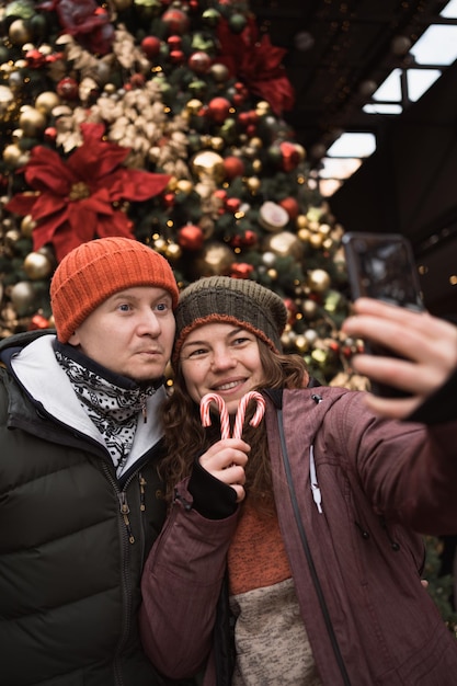 Glückliches Paar mit Hüten und Winterkleidung, das am Weihnachtsbaumhintergrund ein Selfie per Telefon macht