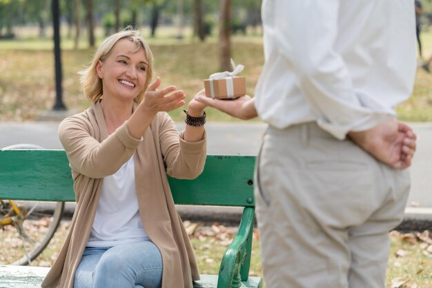 Glückliches Paar Senior Mann Überraschung, die seiner Frau eine Geschenkbox gibt, während sie auf der Bank im Park sitzt