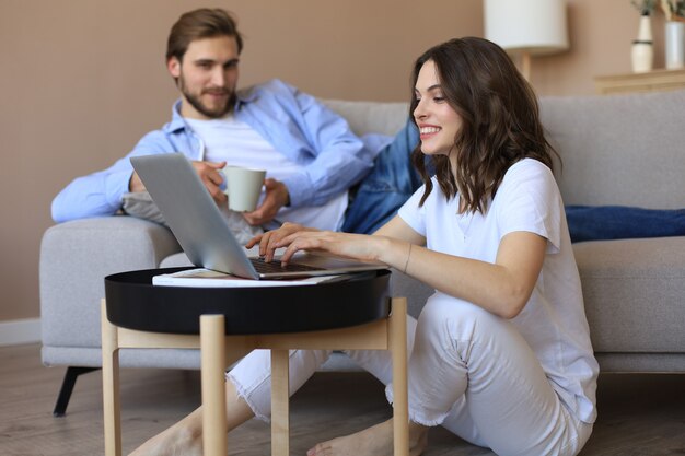Glückliches Paar sitzt, entspannt auf dem Boden im Wohnzimmer, benutzt Laptop für Online-Shopping zusammen, schaut sich einen Film an.