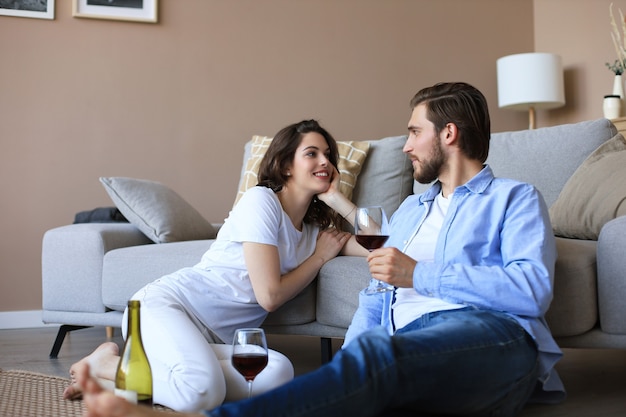 Glückliches Paar sitzt, entspannt auf dem Boden im Wohnzimmer und trinkt Rotwein. Lächelnder junger Mann und Frau ruhen sich zu Hause aus und genießen ein romantisches Date am Familienwochenende zusammen.