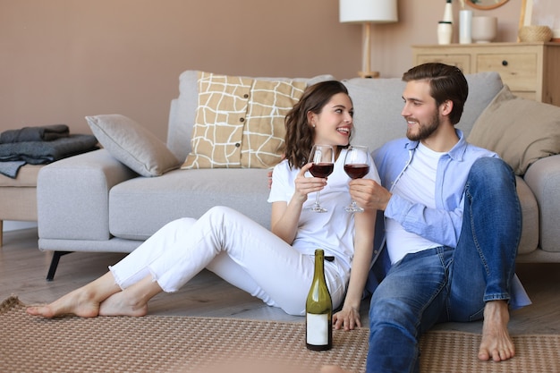 Glückliches Paar sitzt, entspannt auf dem Boden im Wohnzimmer und trinkt Rotwein. Lächelnder junger Mann und Frau ruhen sich zu Hause aus und genießen ein romantisches Date am Familienwochenende zusammen.