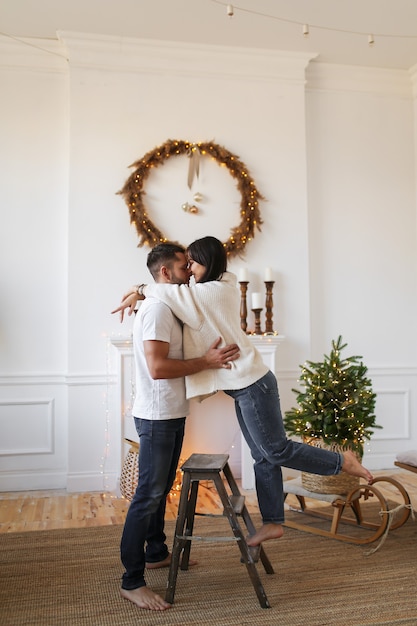 Glückliches Paar verliebt in warmen Pullovern umarmt, küsst und hat Spaß am Kamin und am Weihnachtsbaum
