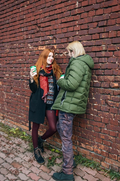 Glückliches Paar von Touristen in der warmen städtischen Kleidung, die mit Kaffee-Einwegbechern auf Ziegelmauer steht