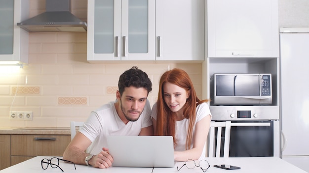 Glückliches Paar wählen, wohin man in den Urlaub fährt, und schaut sich einen Laptop an, der zu Hause in der Küche sitzt