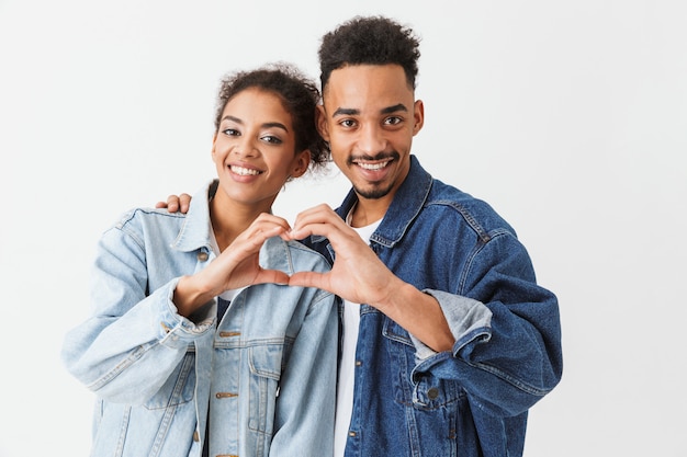 Glückliches reizendes afrikanisches Paar in Jeanshemden, die Herzzeichen über grauer Wand zeigen