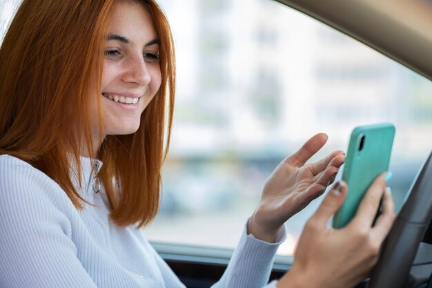 Glückliches rothaariges Mädchen, das auf ihrem Handy hinter dem Lenkrad schreibt, das ein Auto fährt.