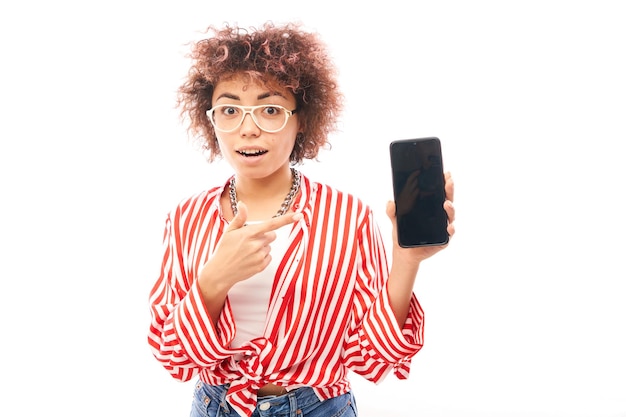Glückliches schockiertes lockiges kaukasisches Mädchen zeigt mit dem Finger auf das Smartphone mit leerem Bildschirm, das auf weißem Studiohintergrund isoliert ist