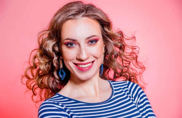 Glückliches schönes brünettes Mädchen mit rotem Make-up, lockigem Haar und einem blauen Streifenpullover, der gegen einen roten Hintergrund aufwirft. Horizontales Foto