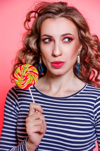 Glückliches schönes brünettes Mädchen mit rotem Make-up, lockigem Haar und einem blauen Streifenpullover, der gegen einen roten Hintergrund aufwirft. Mädchen schaut weg und denkt nach. Vertycal Foto