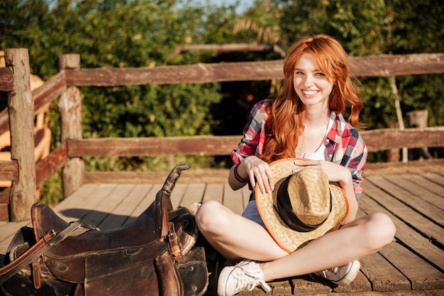 Foto glückliches schönes junges frauengowgirl mit hut, der auf ranch sitzt