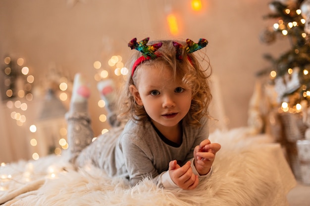 Glückliches schönes kleines Mädchen mit Weihnachtshirschhörnern in modischen Pyjamas, die auf einem Bett vor dem Hintergrund der Lichter liegen