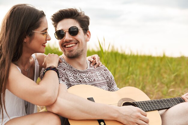 Glückliches schönes Paar, das zusammen im Park sitzt, Gitarre spielt, sich entspannt