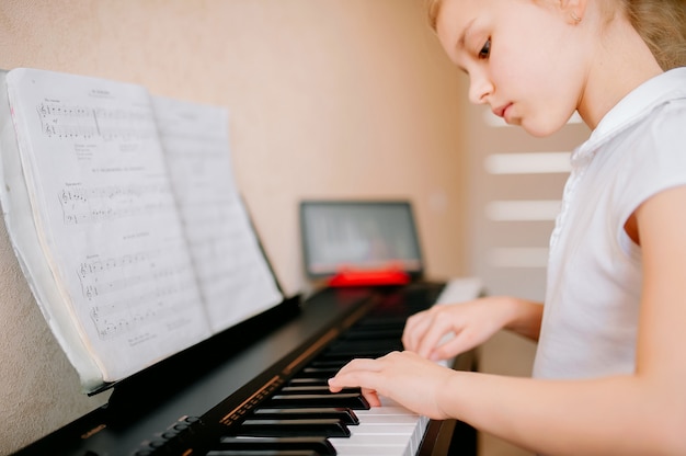 Glückliches Schulmädchen, das Noten studiert und das klassische Digitalpiano spielt, während es eine Online-Lektion auf einem Tablet sieht und lernt, den Synthesizer zu Hause zu spielen, Selbstisolation, Online-Bildung, Fernunterricht