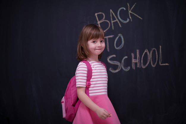 Glückliches Schulmädchen mit Rucksack, das auf schwarzer Tafel zurück zur Schule schreibt