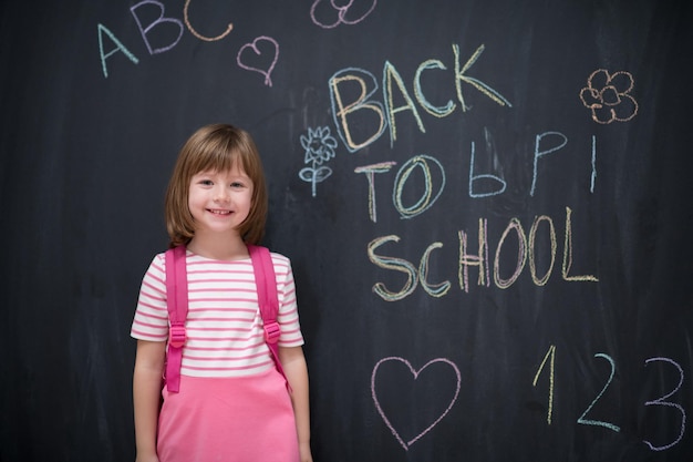 Glückliches Schulmädchen mit Rucksack, das auf schwarzer Tafel zurück zur Schule schreibt