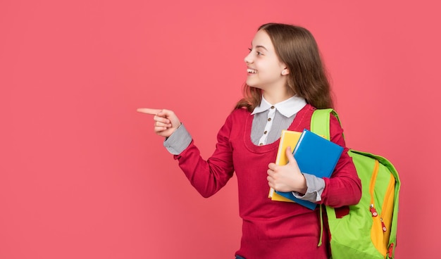 Glückliches Schulmädchen mit Schulbüchern und Tasche, die mit dem Finger für Bildung auf rotem Hintergrund des Kopierraums zeigen