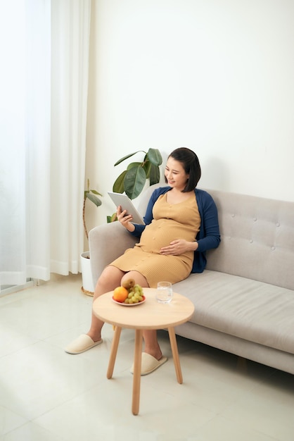 Glückliches schwangeres Mädchen, das im Internet surft und zu Hause Obst isst