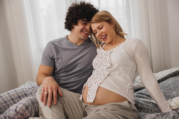 Glückliches schwangeres Paar, das Spaß mit Babykleidung hat, während es sich auf einem Bett im Schlafzimmer entspannt.