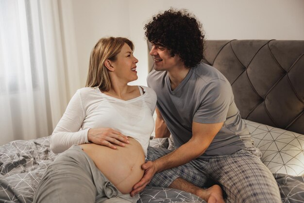 Glückliches schwangeres Paar in Liebe, das kommuniziert, während es sich auf einem Bett im Schlafzimmer entspannt.