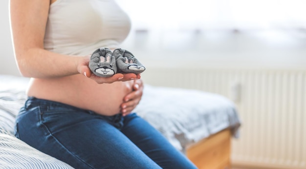 Glückliches Schwangerschaftskonzept Mutter in Blue Jeans hält Babyschuhe Bauch im verschwommenen Hintergrund