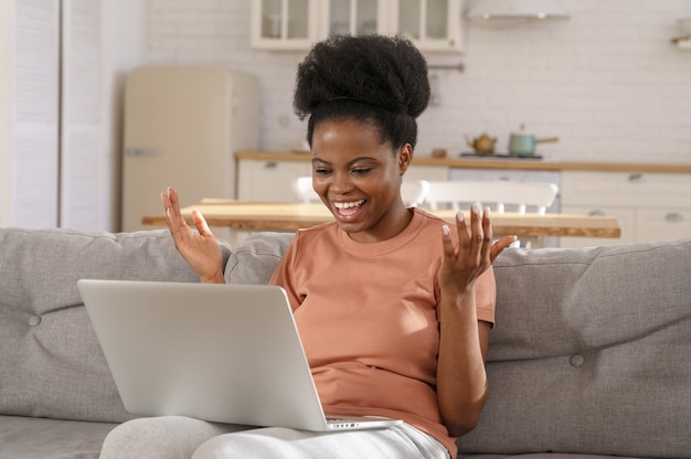 Glückliches schwarzes Mädchen sitzen auf der Couch zu Hause, plaudert mit Freunden per Videoanruf und spricht im Video-Chat.