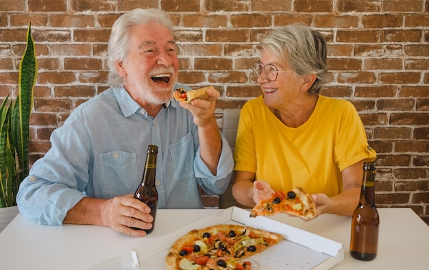 Glückliches Seniorenpaar, das Spaß beim gemeinsamen Essen einer Pizza hat Zu Hause am Tisch mit Bier und Pizza sitzen Feiern Sie Urlaub in Gesellschaft