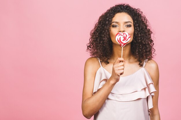 Glückliches sexy amerikanisches Afro-Mädchen, das Lutscher isst. Schönheitsglamourmodellfrau, die rosa süße bunte Lutscherbonbons hält, lokalisiert auf rosa Hintergrund. Süßigkeiten.