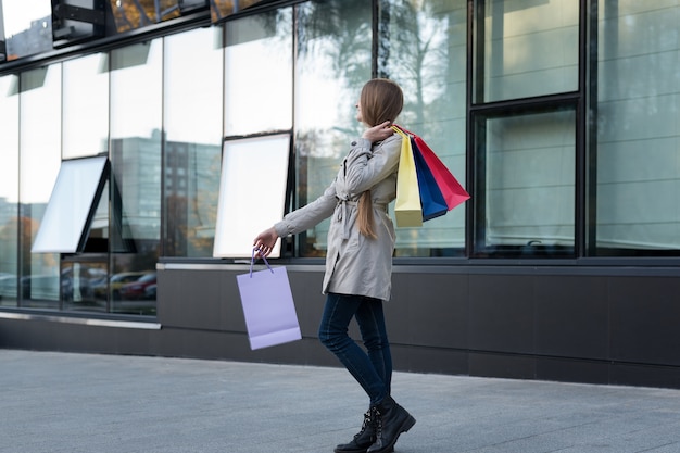 Glückliches shopaholic der jungen Frau mit bunten Taschen nähern sich dem Mall und gehen auf die Straße.