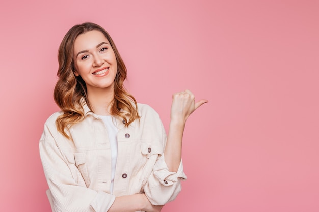 Foto glückliches studentenmädchen lächelt und zeigt mit einem finger auf leer. porträt einer jungen frau lokalisiert über rosa wand