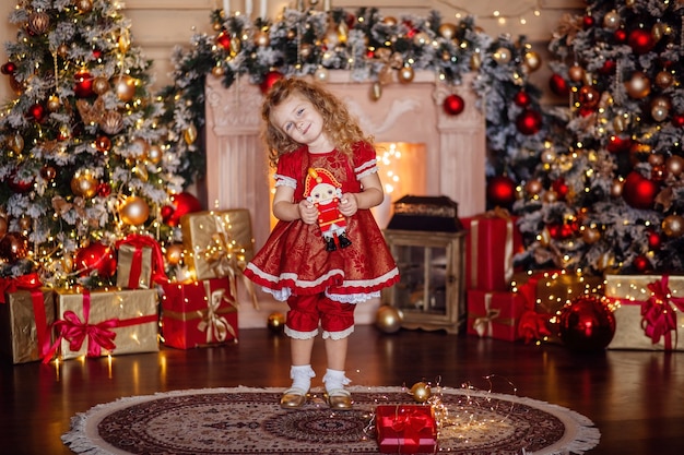 Glückliches süßes kleines blondes Mädchen mit langen Haaren in einem roten Kleid, das auf Weihnachten wartet