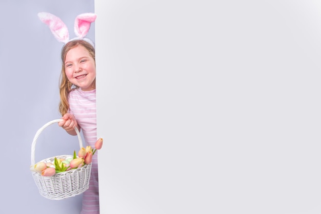 Glückliches süßes Mädchen mit Hasenohren. Ostergrußkartenhintergrund. mit Ostereiern und Frühlingsblume im Korb