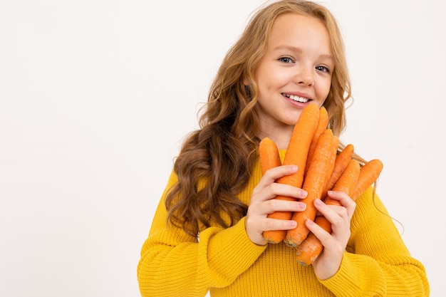 Glückliches Teenager-Mädchen mit roten Haaren, Kapuzenpulli und gelber Hose hält Karotte isoliert auf Weiß