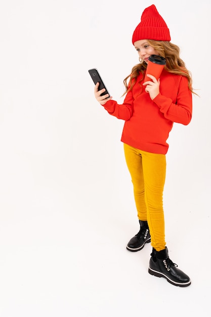 Glückliches Teenager-Mädchen mit roten Haaren, rotem Hut, Kapuzenpulli und gelber Hose lächelt, trinkt Kaffee und spielt Telefonspiele isoliert auf weißem Hintergrund