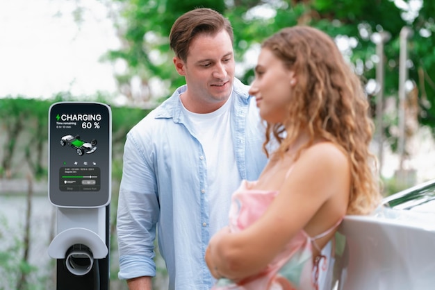 Glückliches und liebenswertes Paar mit umweltfreundlichem, bewusstem Elektrofahrzeug, das von einer Elektrofahrzeug-Ladestation aufgeladen wird. Elektrofahrzeugtechnologie wird als alternative Verkehrsmittel für die zukünftige Nachhaltigkeit genutzt.