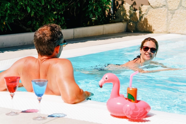 Glückliches und schönes junges Paar, das Freizeit am Pool genießt. Spaß romantischer Lebensstil. Urlaubsreise im Ferienort. Zusammen reisen. Blick auf Erwachsene Menschen entspannen sich im Sommer. Frau schwimmt in der Nähe ihres Mannes