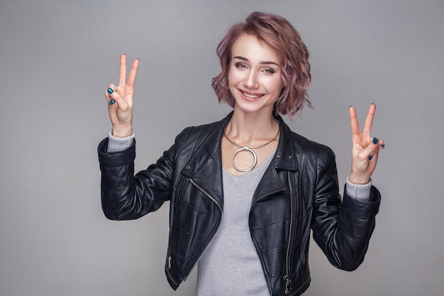 Glückliches zahniges Smiley-schönes Mädchen mit kurzer Frisur, Make-up in lässiger schwarzer Lederjacke, die mit Sieg- oder Friedenszeichen steht und die Kamera ansieht. Studioaufnahme, auf grauem Hintergrund isoliert