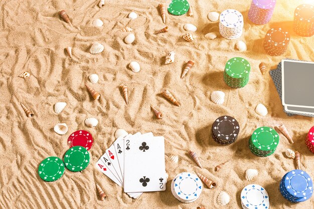 Glücksspiel im Urlaub Konzept weißer Sand mit Muscheln farbige Pokerchips und Karten Draufsicht