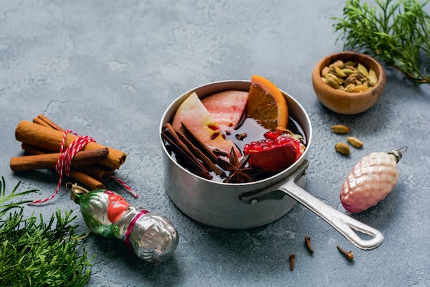 Glühwein-Heißgetränk mit Zitrusfrüchten, Apfel, Granatapfel und Gewürzen in Aluminiumauflauf mit Vintage-Weihnachtsbaumspielzeug und Tannenzweig auf Betonoberfläche.