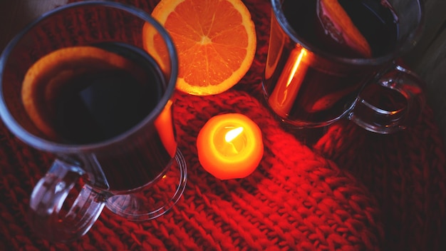 Glühwein in Glasbechern, brennende Kerze auf dunkelrotem Hintergrund. Glühwein mit orangefarbenem und rotem Schal