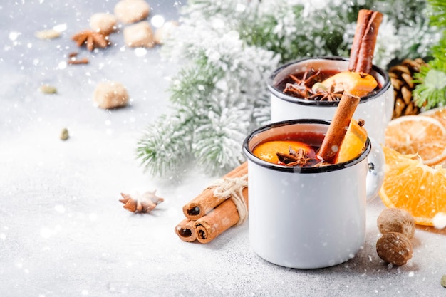 Glühwein in Weißmetallbechern mit Zimtgewürzen und Orange mit Tanne und Weihnachtsbeleuchtung traditionelles Getränk im Winterurlaub Kopieren Sie Platz für Text