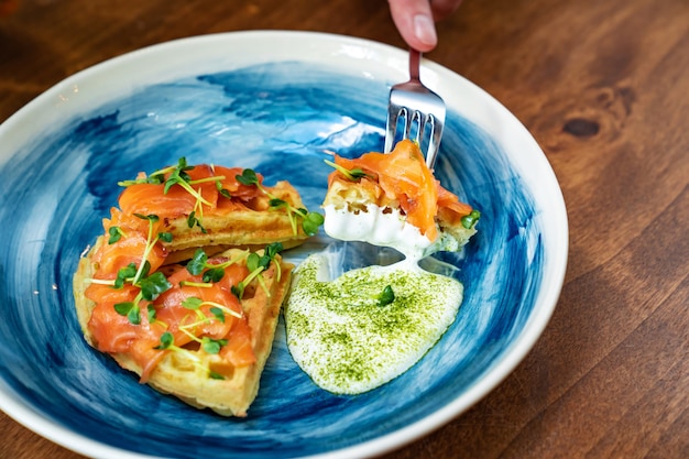 El gofre vienés con salmón y salsa blanca en un plato azul se come con cuchillo y tenedor.