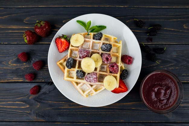 Gofres belgas con bayas de verano y azúcar en polvo en un plato blanco sobre un fondo de madera oscura Gofres belgas dulces para el desayuno o el almuerzo