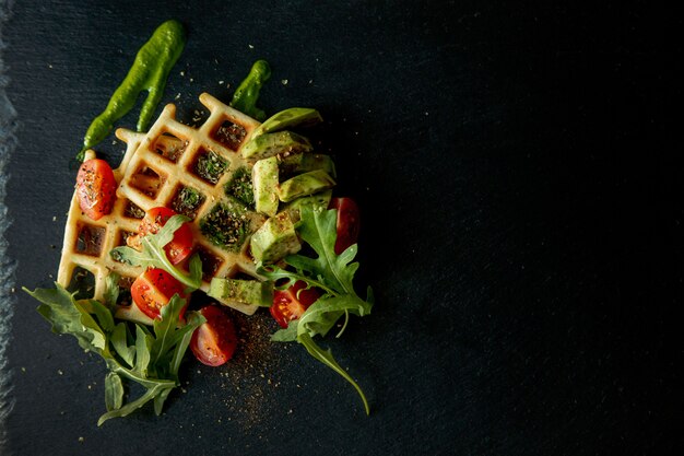 Gofres belgas recién horneados con rúcula, tomate y aguacate en un plato negro. Gofres salados. Concepto de desayuno. Desayuno saludable