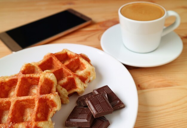 Gofres belgas y trozos de chocolate negro con café caliente borroso y teléfono inteligente en segundo plano.