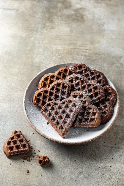 Gofres de chocolate en forma de corazón espolvoreados con azúcar en polvo en un plato sobre fondo texturizado. Concepto para el día de san valentín