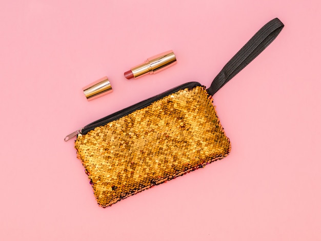 Goldene Damenhandtasche mit Lippenstift auf einem rosa Tisch